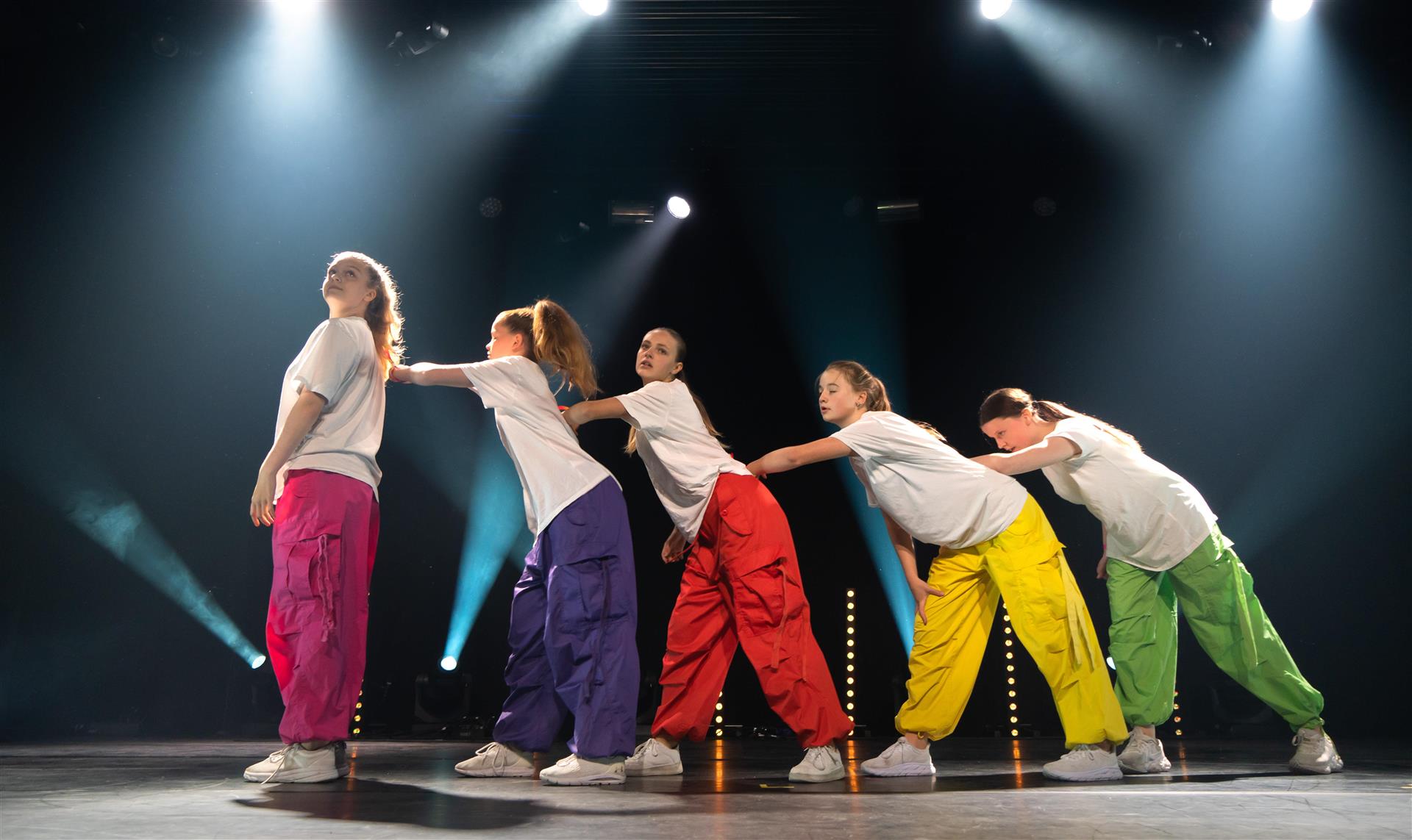 Fem jenter i fargerike bukser danser på ei scene.