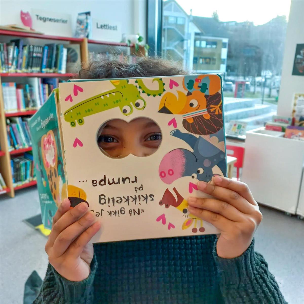 Barn med bok framfor ansiktet. - Klikk for stort bilete